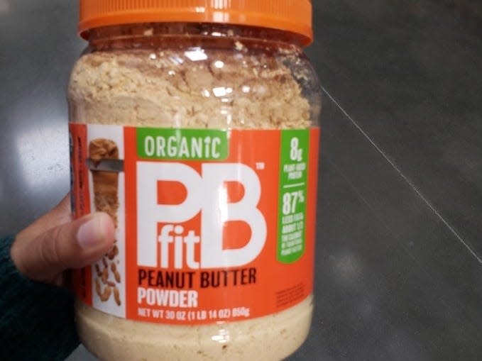 Hand holding clear and orange container of PBFit peanut butter powder at costco