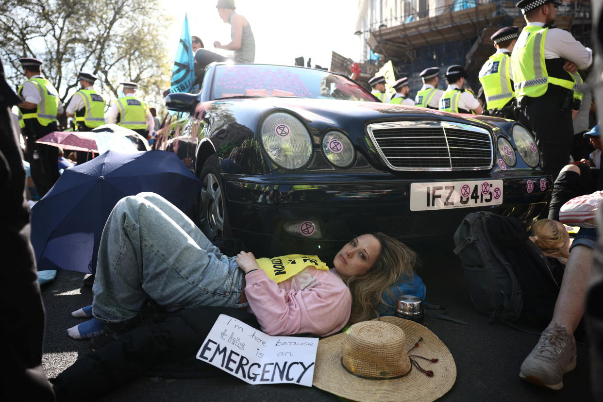 A female activist from the Extinction Rebellion lies in front of a car with her straw hat on the ground and a poster lying by her saying: I'm here because this is an Emergency. Two rows of London police officers in lime-green harnesses stand parallel to the vehicle. 