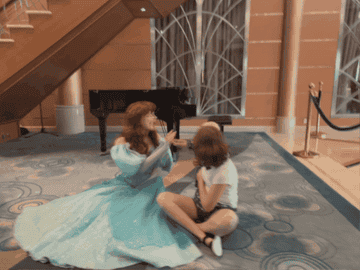 Merida from Disney's Brave animated movie sits on the floor next to a young fan, sharing a moment under a staircase indoors