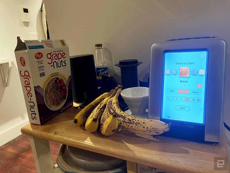 The Revolution InstaGLO R180 Toaster on a countertop next to a bunch of bananas, a pour-over coffee maker and a box of Grape Nuts cereal.