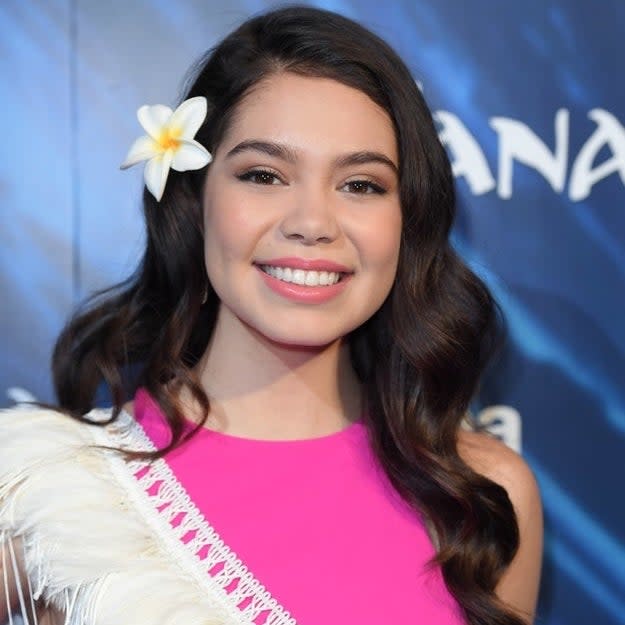 Aulii Cravalho smiles in a pink dress with white fringe detail and a flower in her hair