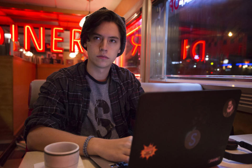 Cole Sprouse in a casual outfit sits at a booth in a diner working on a laptop, with neon lights in the background