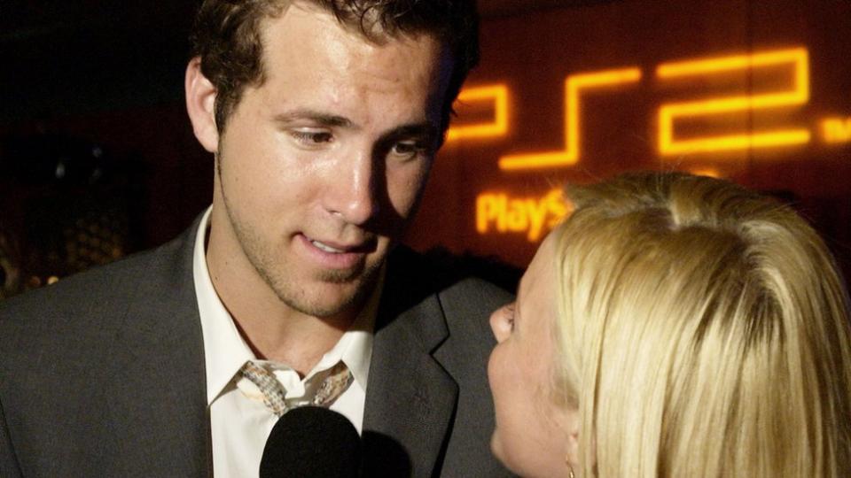 Takım elbiseli genç Ryan Reynolds, soru sorarken görüşmeciye bakıyor.  İkisi de Playstation 2 logosunun turuncu harflerle gösterildiği bir pankartın önünde duruyor.