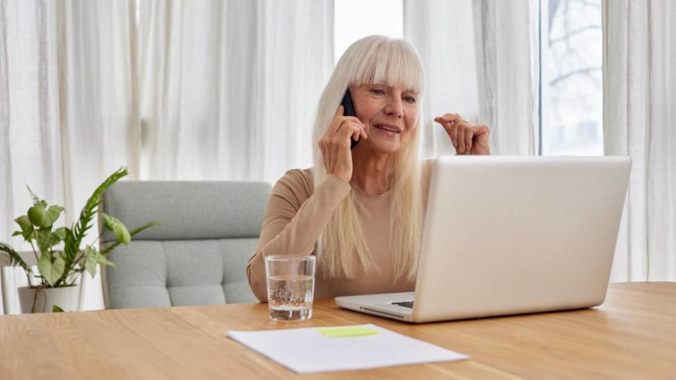 IRS work from home jobs: Elderly woman working on laptop computer, smiling, talking on the phone. Senior woman using laptop. Elderly woman sitting at home, using laptop computer and talking on her mobile phone, smiling