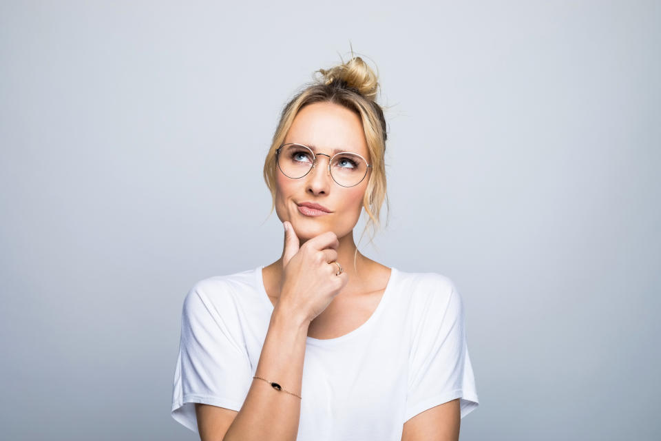 Woman in a white shirt and glasses with a thoughtful expression, hand on chin, hair in a top knot