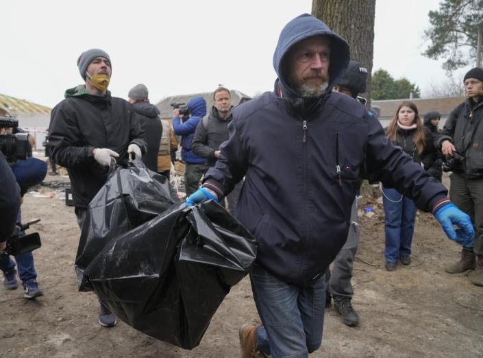 A man, right, wearing a hooded jacket and blue gloves carries a black body bag with the help of another man