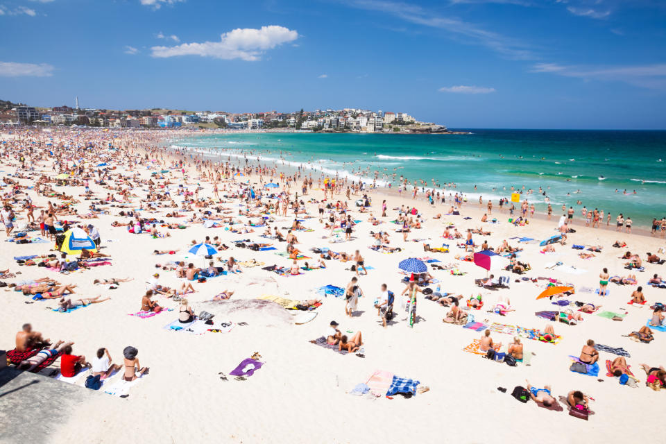 Crowded beach scene with people sunbathing, swimming, and relaxing under umbrellas. In the background, buildings and greenery are visible along the coastline