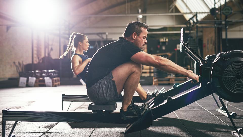 Man and woman on a rower in a warehouse gym, man is reaching forward to grip the handlebars of his rower