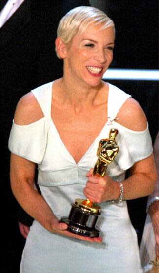 Singer Annie Lennox is wearing a white dress and smiling as she holds her Oscar