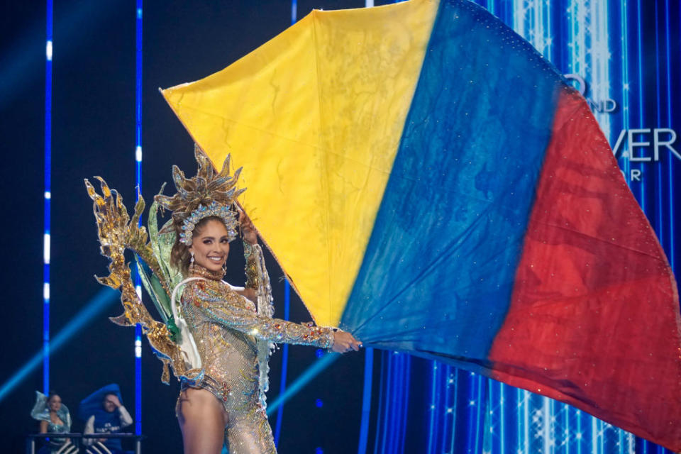 She's carrying the colors of the flag — blue, red, and gold — and wearing an ornate, long-sleeved tasseled, bejeweled bodysuit with ornate headdress