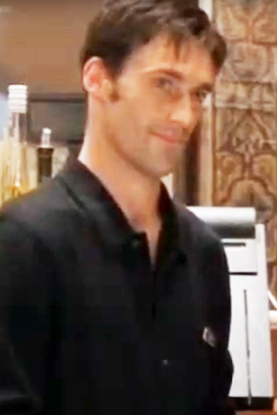 A young Jon Hamm in a black collared shirt stands in front of a bar setup, smiling slightly