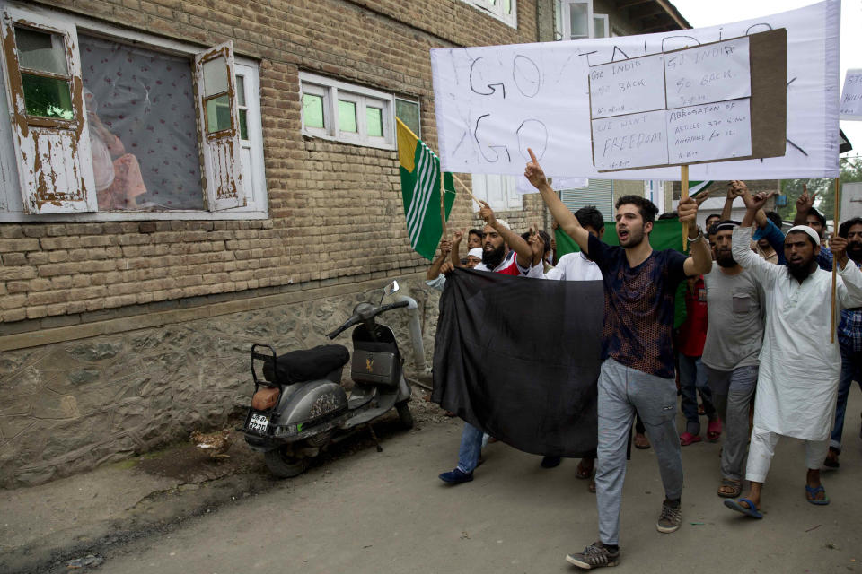 A Kashmiri Muslim woman, left, watches from her house as men hold placards and shout pro-freedom slogans during a demonstration after Friday prayers amid curfew like restrictions in Srinagar, India, Friday, Aug. 16, 2019. Hundreds of people have held a street protest in Indian-controlled Kashmir as India's government assured the Supreme Court that the situation in the disputed region is being reviewed daily and unprecedented security restrictions will be removed over the next few days. (AP Photo/Dar Yasin)