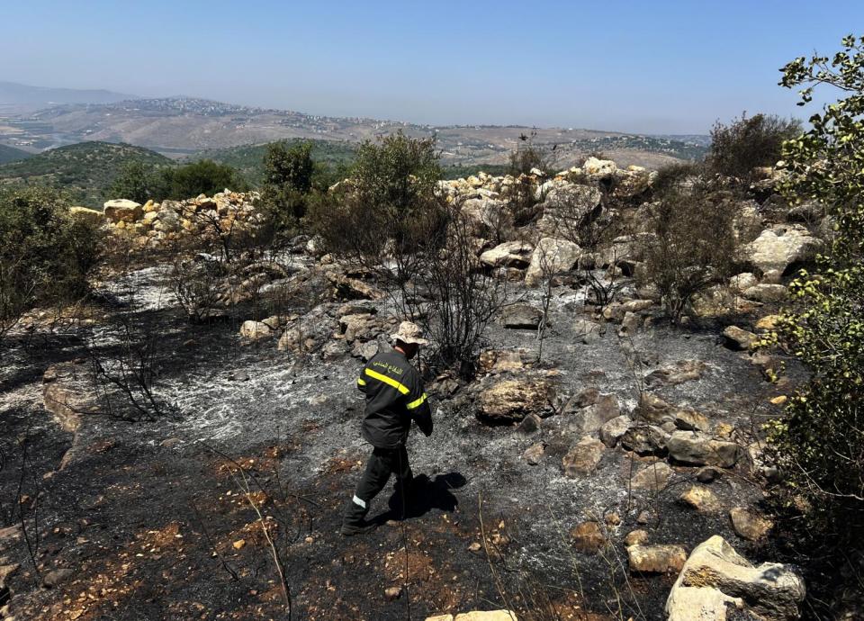 A man wearing a jacket with yellow reflective strips trudges through a burned, soot-covered meadow.