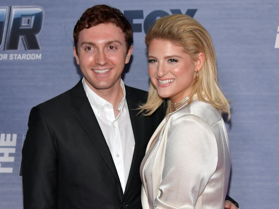 daryl sabara and meghan trainor a red carpet, posing together and smiling. trainor is wearing a silken white dress, and sabara is wearing a black suit