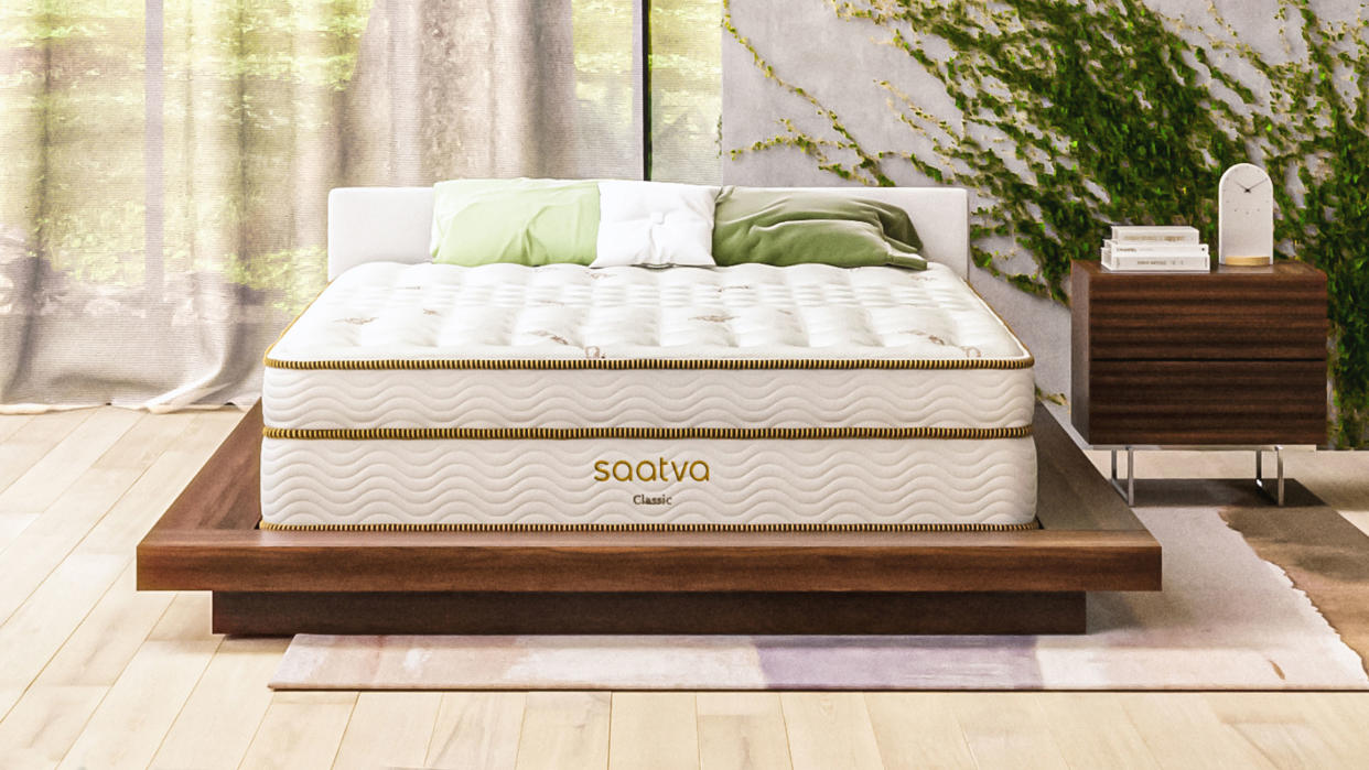  Saatva Classic Mattress review image shows the Saatva Classic innerspring hybrid placed on a luxury wooden bed frame and dressed with white and green pillows. 