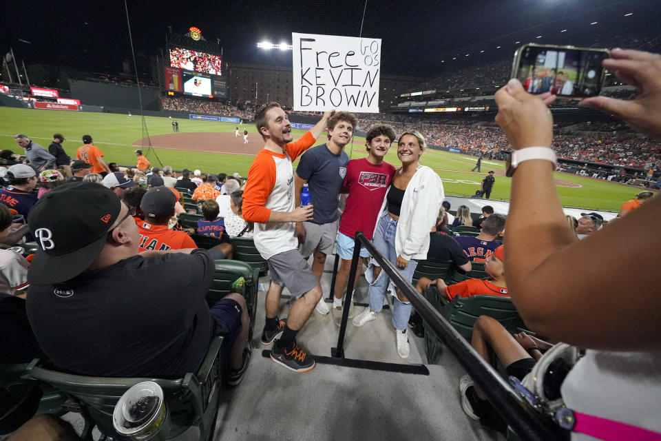 Kevin Tehansky, left, holds a sign in support of broadcaster Kevin Brown while photo bombing a group of people posing for pictures at Oriole Park at Camden Yards in the eighth inning of a baseball game between the Baltimore Orioles and the Houston Astros, Tuesday, Aug. 8, 2023, in Baltimore. The Astros won 7-6. The Orioles indicated that Brown would be back on the air soon after reports that his recent absence was because he mentioned that the team had already won as many games at Tampa Bay this year as it did over the previous three seasons. Announcers such as Michael Kay of the YES Network came to Brown's defense. The Athletic, citing unidentified sources, reported that Brown was taken off the air over his comments on a MASN-TV broadcast July 23. A team official says the Orioles don't comment on personnel matters but were looking forward to having Brown back soon. (AP Photo/Julio Cortez)