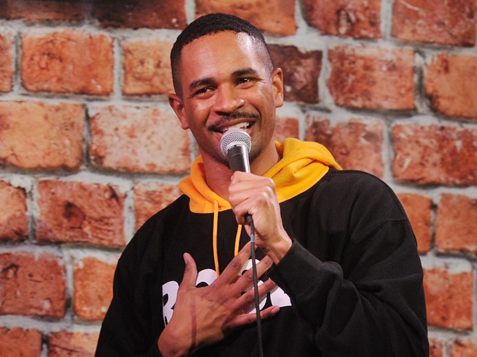 Damon Wayans Jr. in a black sweatshirt with a yellow hood holding a microphone next to a sign that says "Factory Comedy Club"