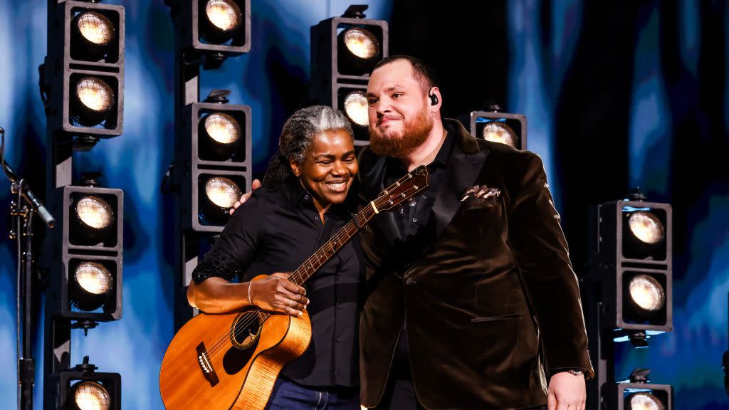 tracy chapman and luke combs hug and smile while standing on a stage in front of several lights, she holds an acoustic guitar