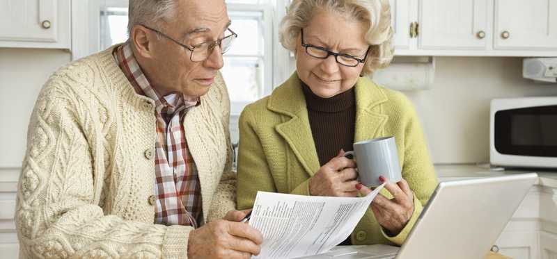 An elderly couple reviews paperwork with laptop in front of them.