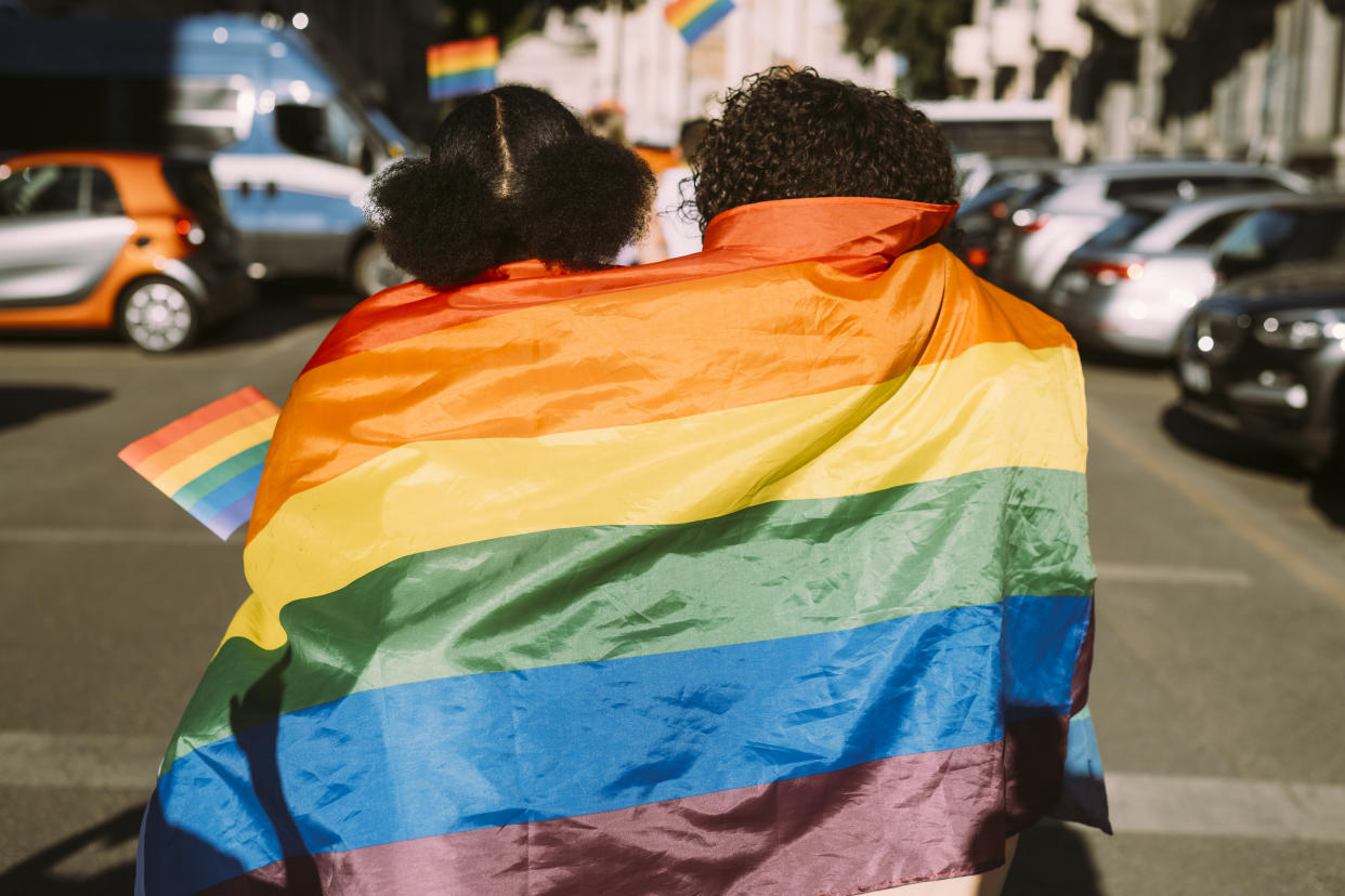 Two young people walk together down the street with a pride flag draped around their shoulders.