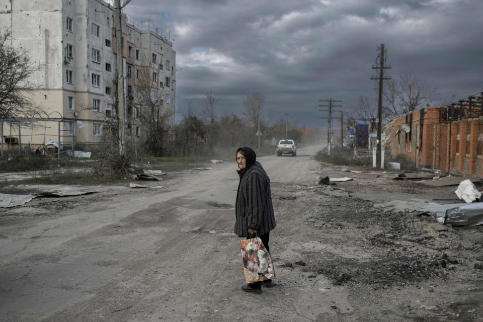 An old woman in a scarf carrying shopping bags looks disconsolately down a rutted dirt road lined with a dilapidated apartment building. A pickup drives away in the distance.