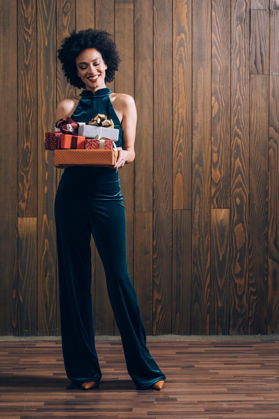 A woman in a velvet jumpsuit holds a stack of presents.