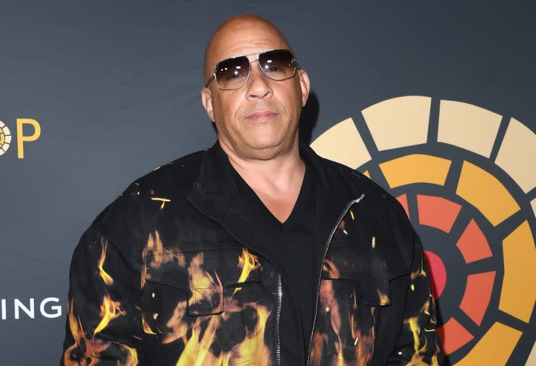 Vin Diesel, wearing a flame-patterned jacket and sunglasses, stands in front of a backdrop at a work-related event