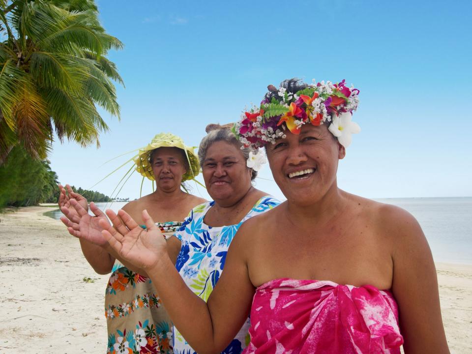 Three women in brightly colored clothing and headware stand and wave on a beach on Cook Island