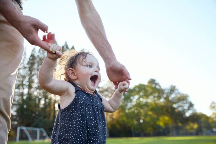 Toddler walking with adult's support, expressing joy, outdoors in a park setting