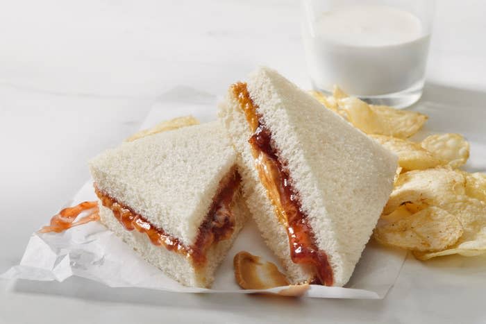 Peanut butter and jelly sandwich with a bite taken, next to potato chips and a glass of milk