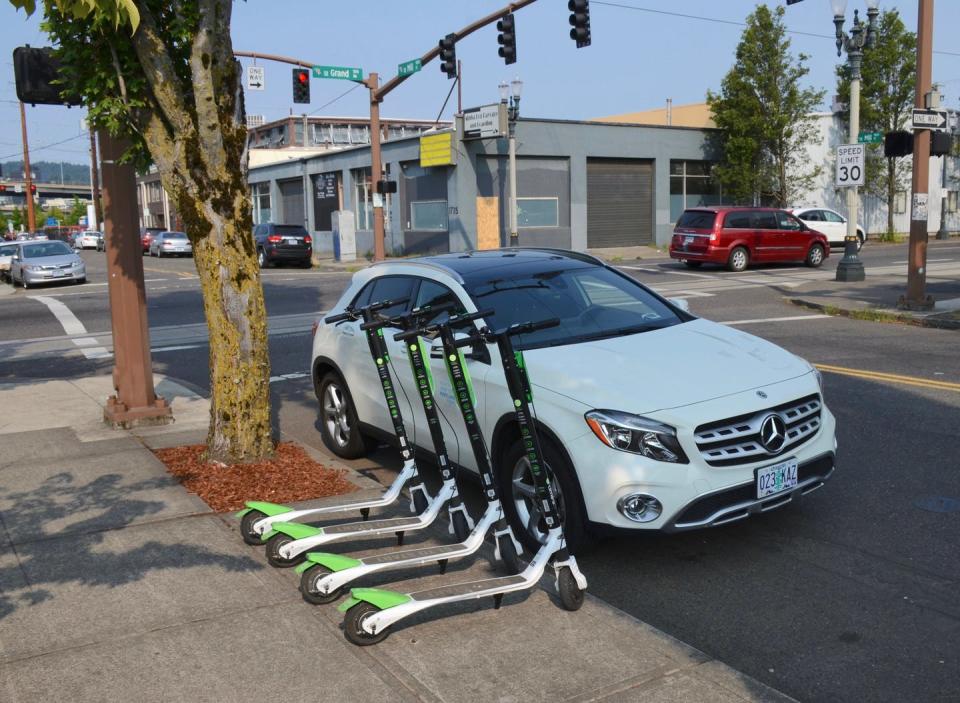 Four scooters lined up on the edge of a sidewalk, near the street and a car
