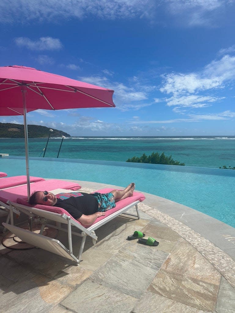 A man laying down on a pool chaise lounge with a pink cushion and pink umbrella next to an infinity pool.