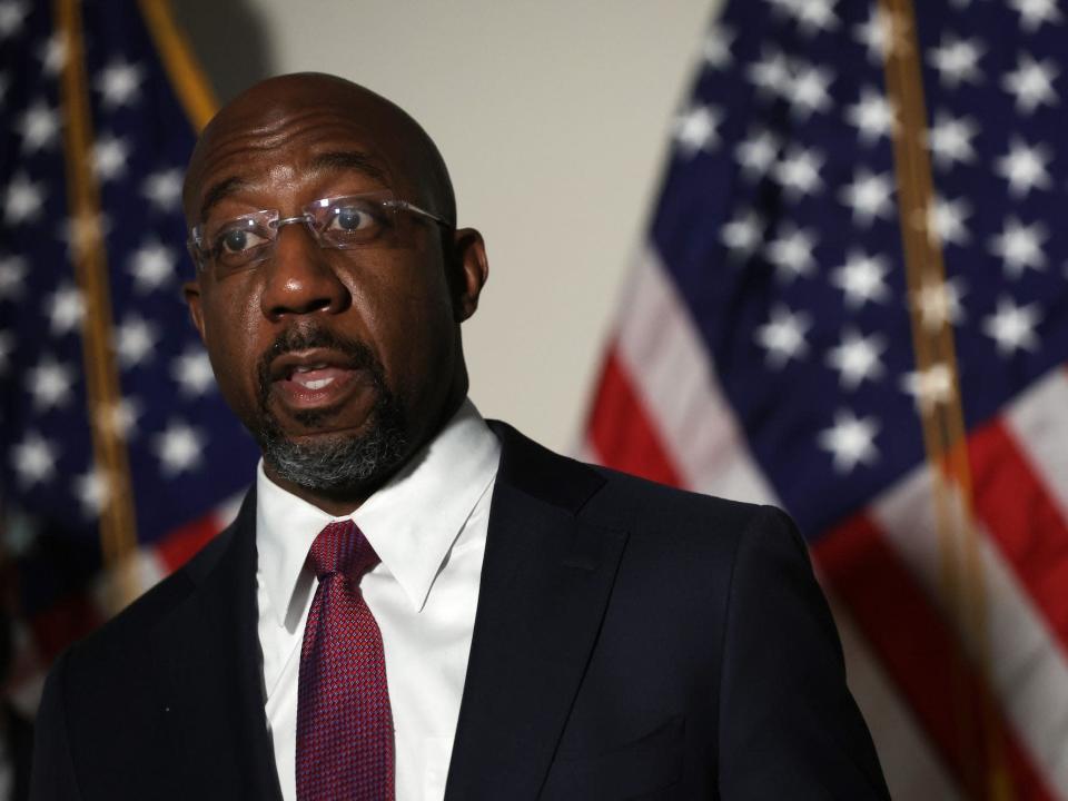 Suit-clad Democratic Sen. Raphael Warnock of Georgia speaks to reporters while standing against a backdrop of American flags at the US Capitol.
