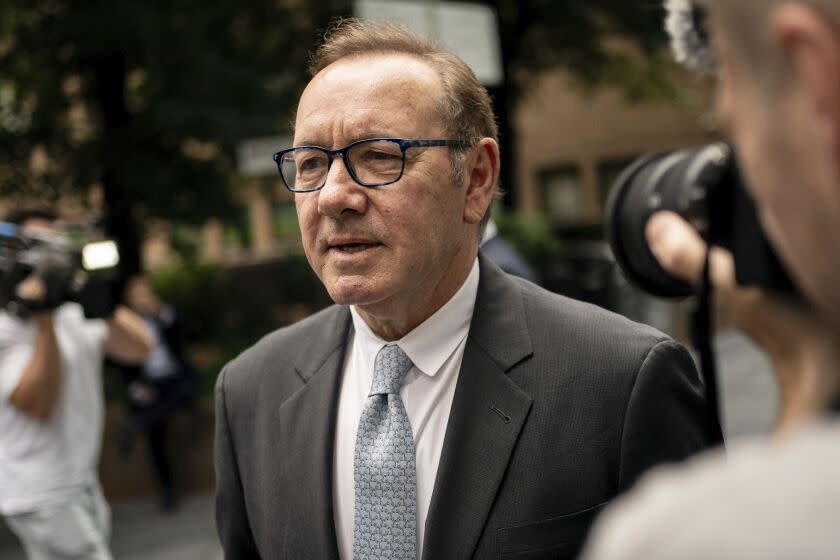 Kevin Spacey is wearing a gray suit with black thick-framed glasses while walking into courthouse, being photographed