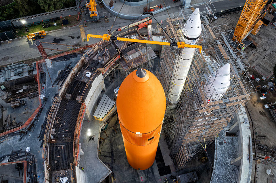 Aerial view of a large orange cylindrical fuel tank and two white rockets, surrounded by scaffolding.
