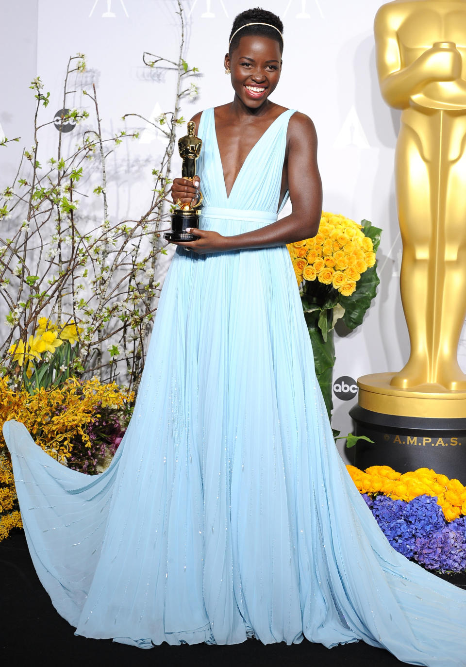 Lupita Nyong'o holding an Oscar, wearing a plunging V-neck light blue gown with a flowing skirt, standing beside a golden statue