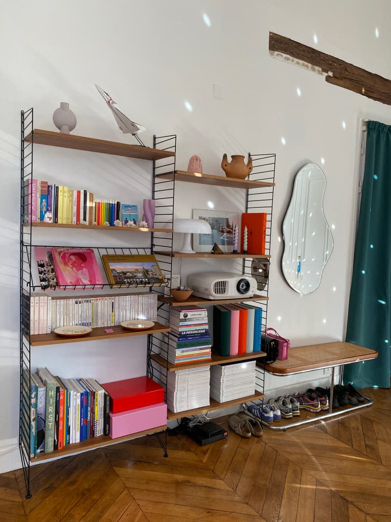 Eclectic bookshelf in apartment with wavy mirror above caned bench.