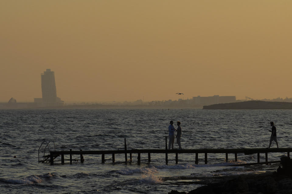 People walk on a sea deck as in the background is seen an apartments tower of a new construction of "Ayia Napa Marina", during sunset in the resort of Ayia Napa in the eastern Mediterranean island of Cyprus, on Tuesday, June 6, 2023. When the U.S. and U.K. in April included a handful of Cypriot nationals and Cyprus-registered companies as part of another global crackdown on 'enablers' helping Russian oligarchs skirt sanctions, the perception that the island nation somehow remains Moscow's financial lackey again loomed large. (AP Photo/Petros Karadjias)