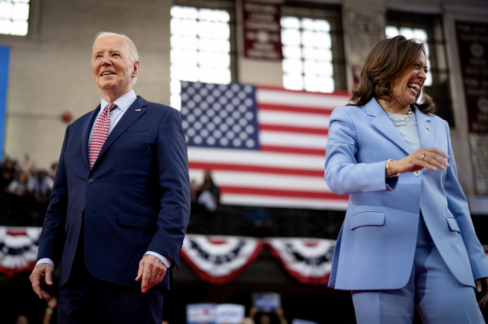 Joe Biden and Kamala Harris stand indoors, smiling and wearing suits, with a large American flag behind them