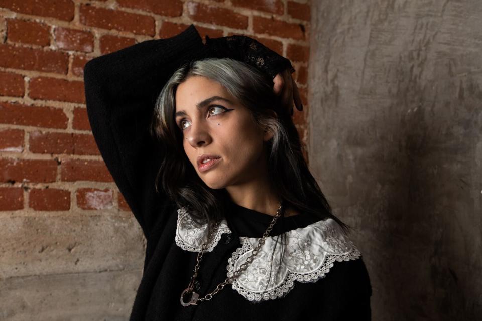 A woman wraps an arm atop head in front of an exposed brick wall.