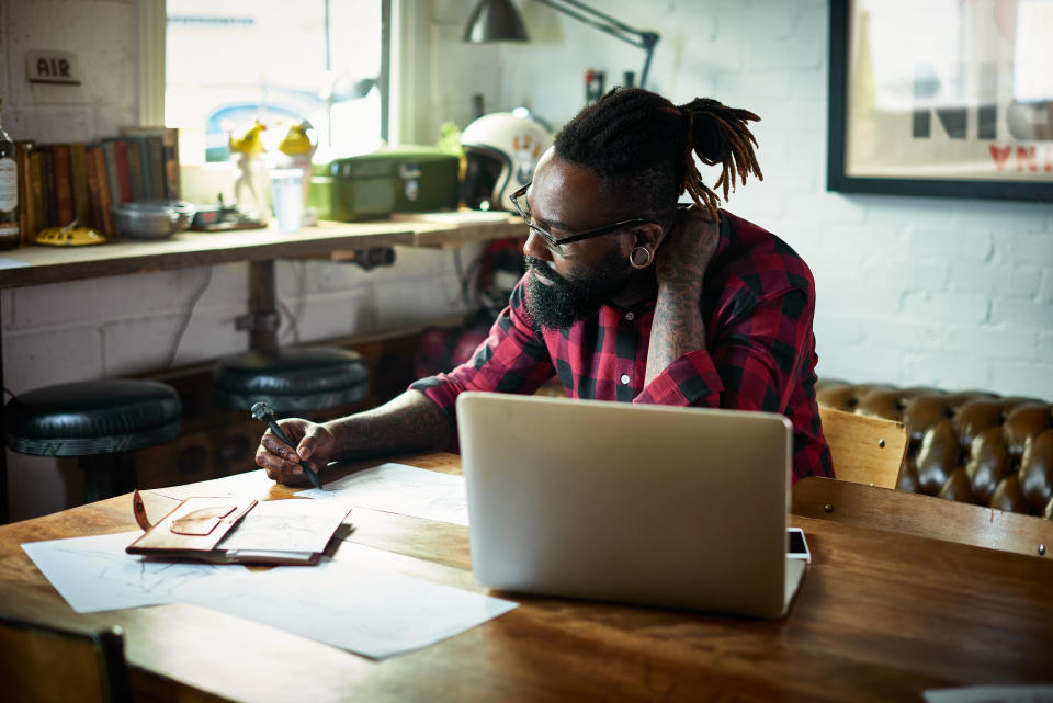 Bearded designer with tattoos drawing at desk in front of laptop.