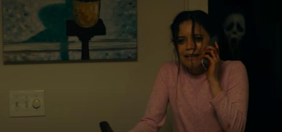 Tara holding a knife with a phone to her ear while Ghostface runs up behind her from the dark in "Scream" (2022)