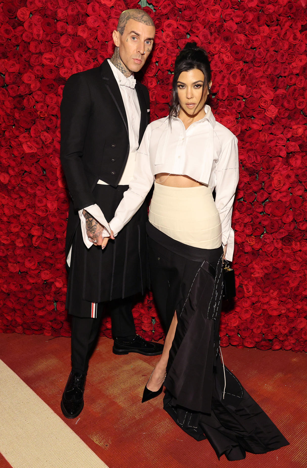 Travis Barker and Kourtney Kardashian hold hands and pose in front of a wall of red roses at the Met Gala