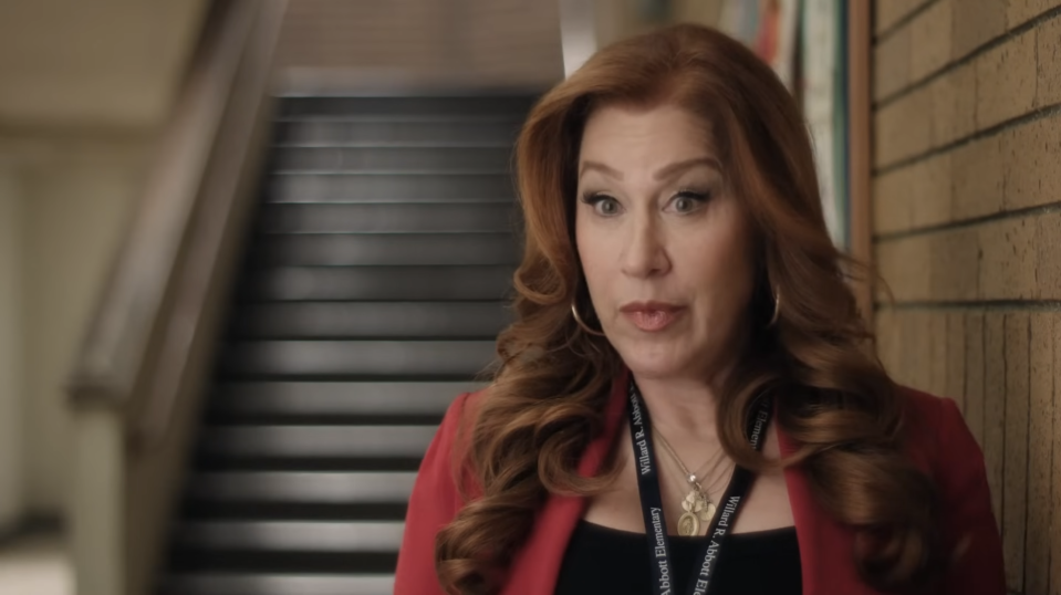 Woman in a red blazer and necklace speaks in a school hallway setting