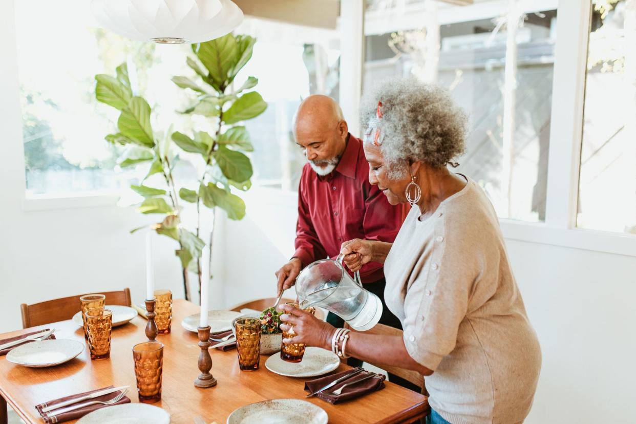 Senior woman pouring water in glass from jug at home. Retired couple is arranging dining table in room. They are preparing together.