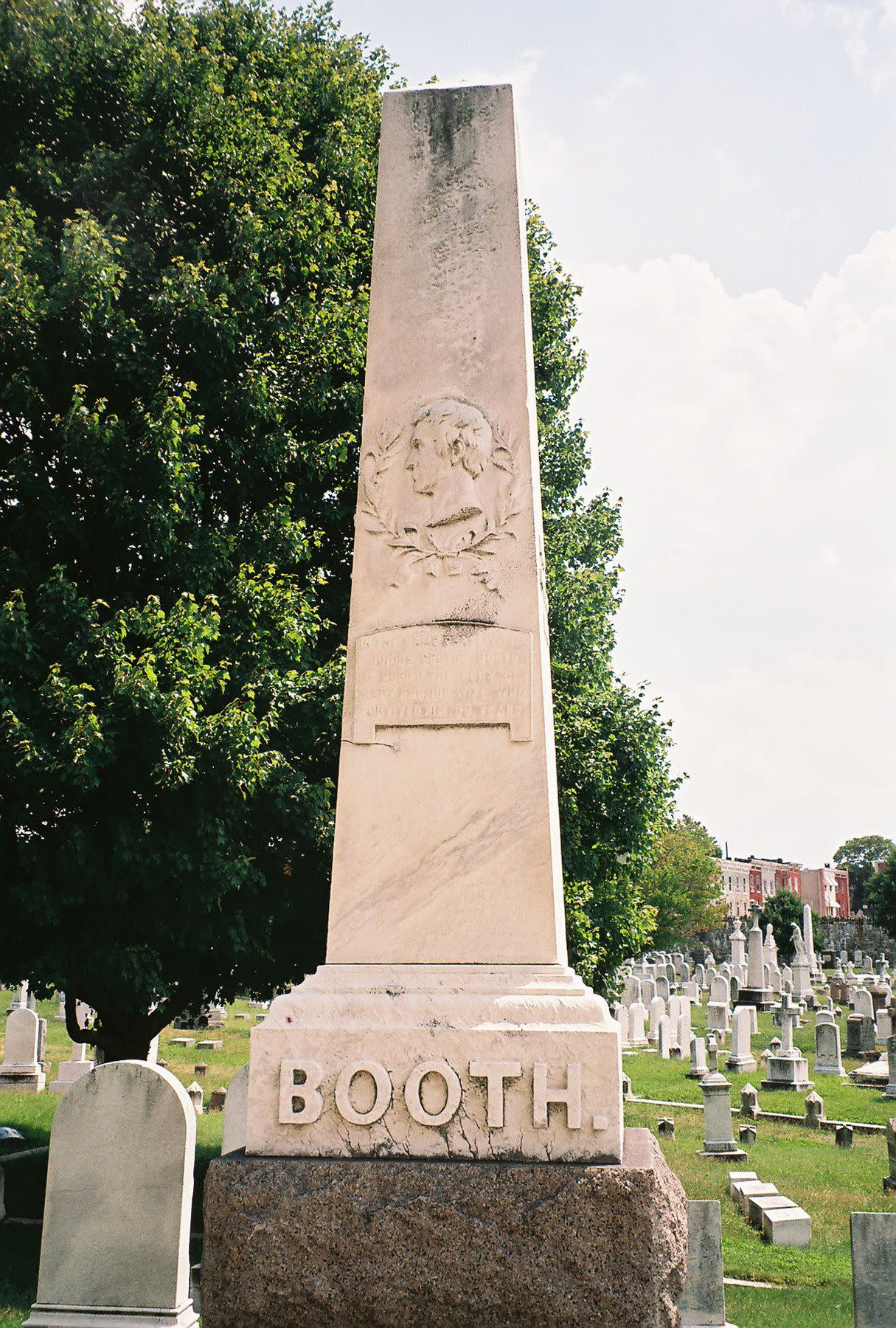 John Wilkes Booth's Grave in the Greenmount Cemetery, Baltimore, a tall monument among several other graves, with a big tree to the left on a sunny summer day