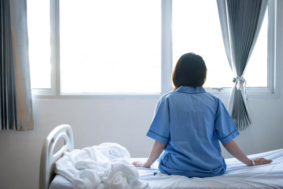 A person in a hospital gown sits on a bed facing away from the camera, looking out a window with open curtains. The bed is unmade