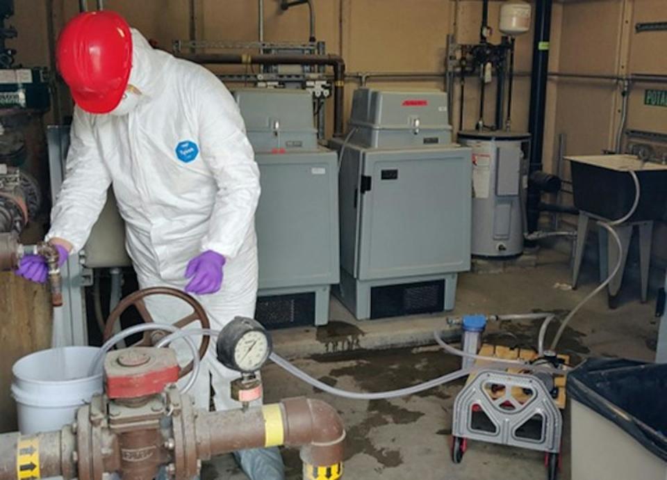 person covered in protective gear and hard hat runs water into a bucket in industrial setting