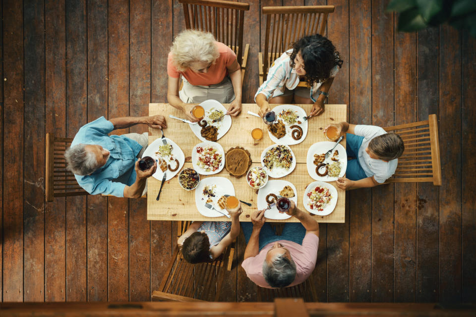 Aerial view of six people of various ages and genders seated around a wooden table sharing a meal. The table is laden with various dishes and beverages