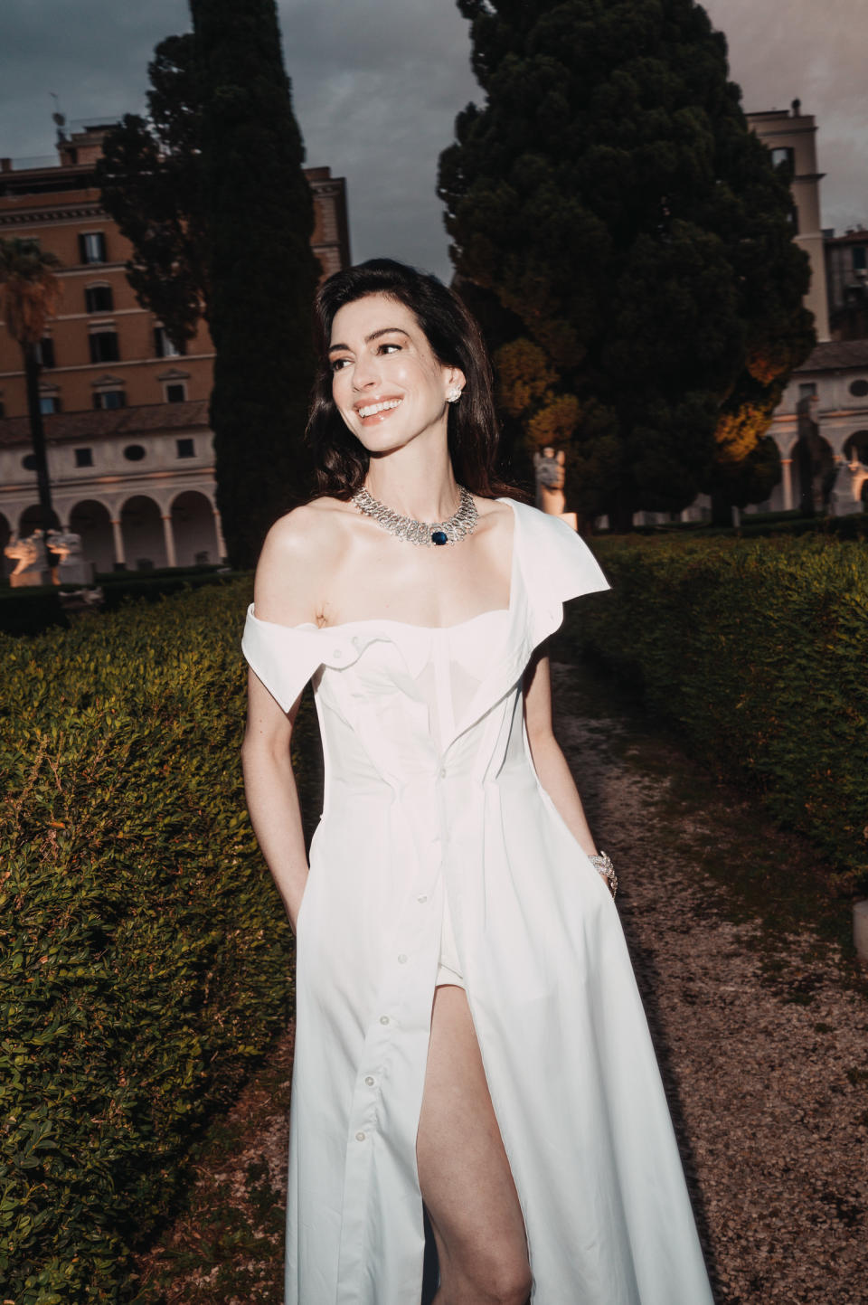 Anne Hathaway poses in an elegant off-the-shoulder gown with a high slit, standing outside with hedges and trees in the background